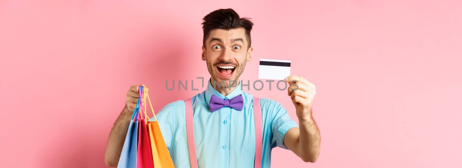 Happy young man giving his credit card and holding shopping bags, buying with discounts, standing on pink background and smiling.