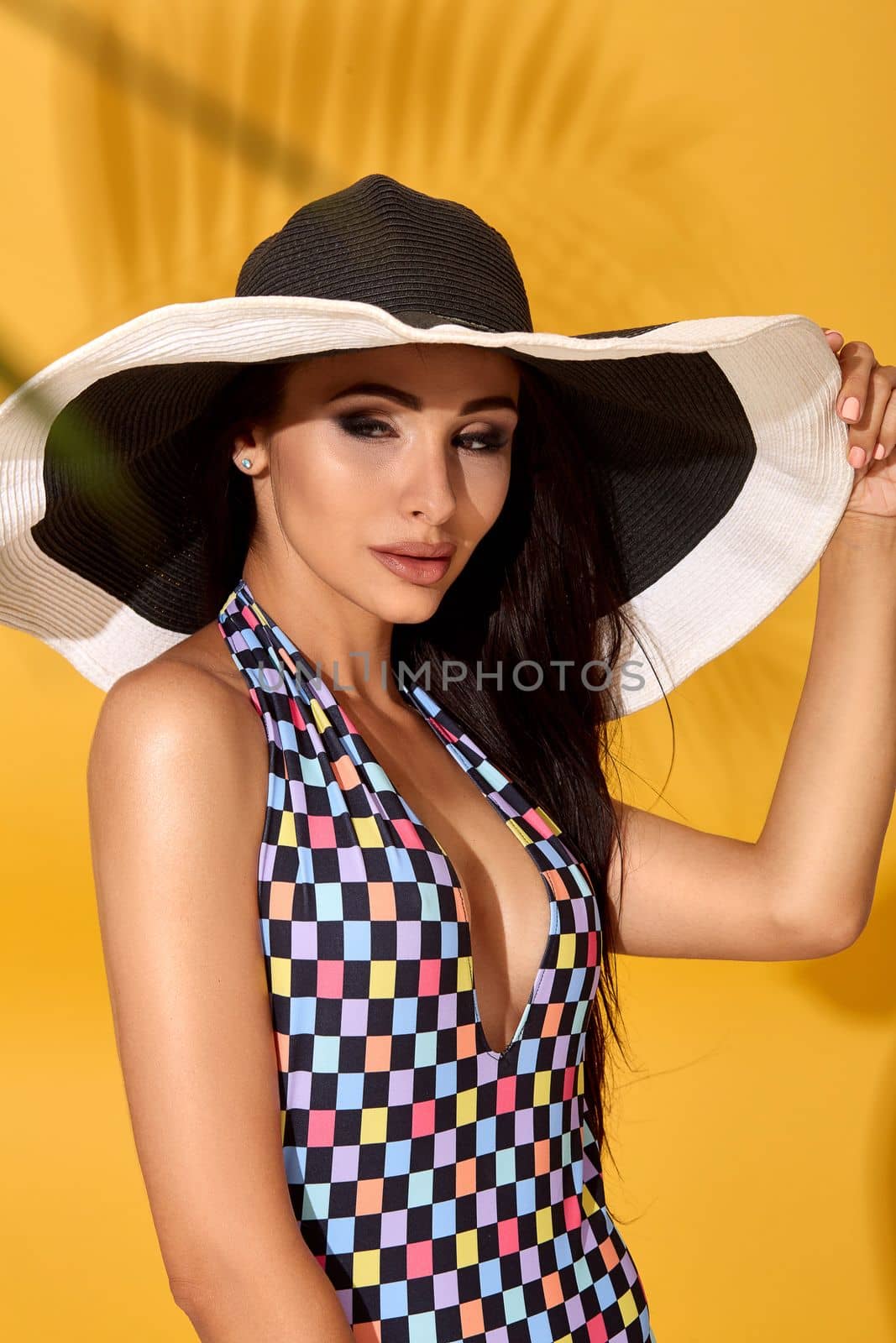 Photo of young beautiful brunette girl posing in the studio, standing in a black-and-white hat and checkered swimsuit. Yellow background