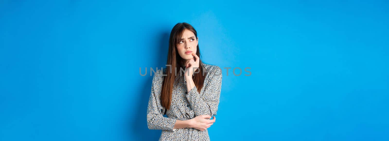 Thoughtful serious woman in dress looking away pensive, standing hesitant and unsure, thinking on blue background.