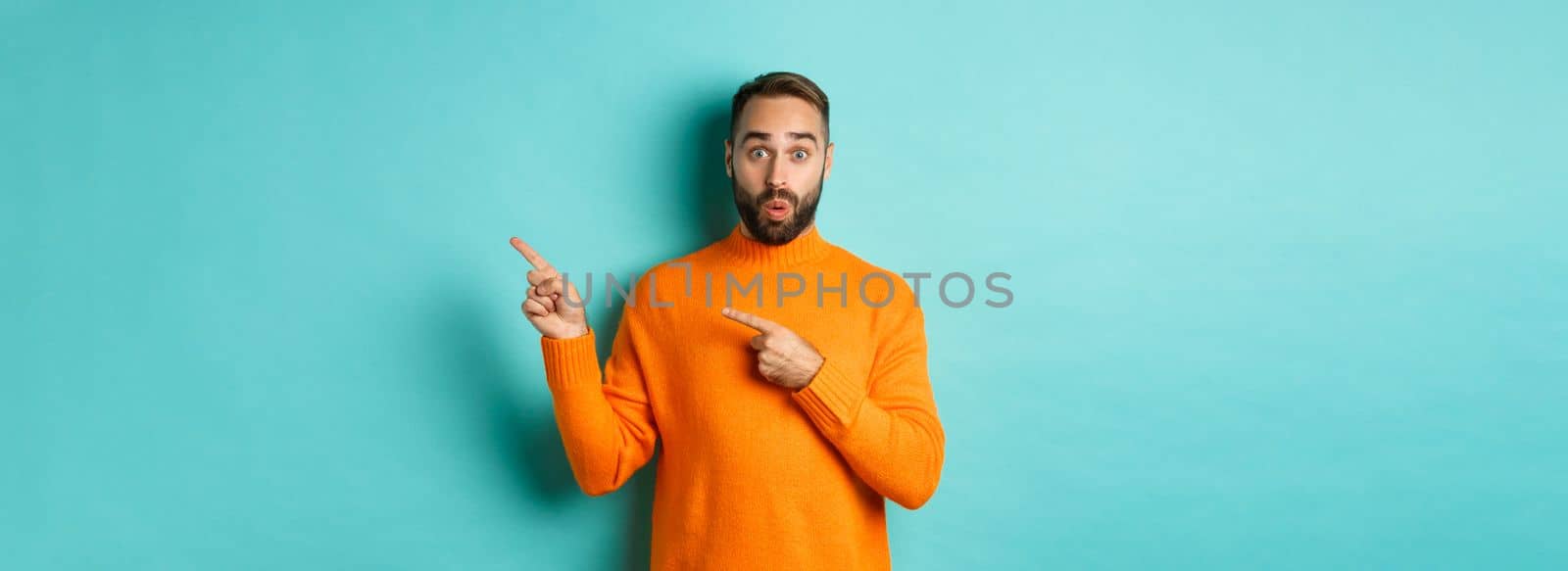 Amazed guy showing advertisement, pointing fingers right at banner, standing against turquoise background.