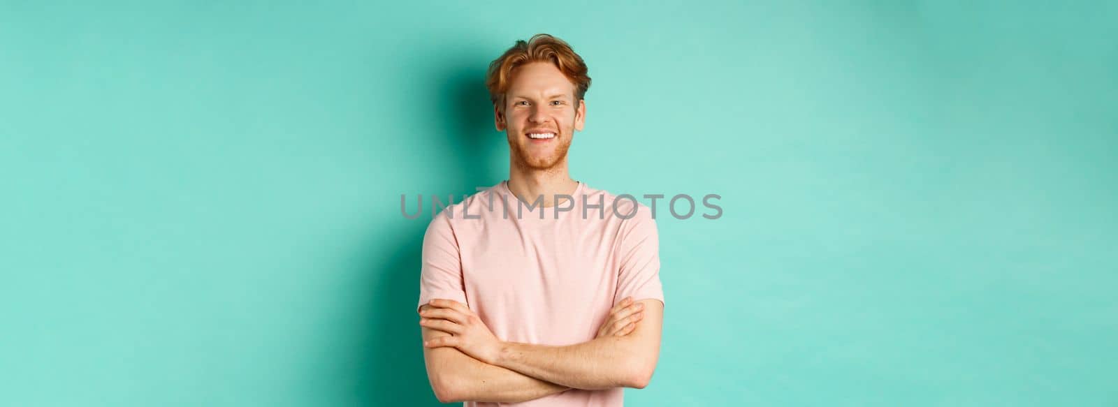 Portrait of friendly-looking young man with red hair and beard smiling and starng satisfied, holding hands crossed on chest, standing over turquoise background.