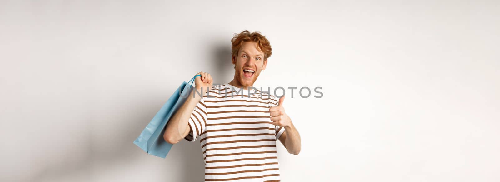 Happy young man with red hair, shopping in stores, showing thumbs-up and holding paper bag over shoulder, recommending shop, white background by Benzoix