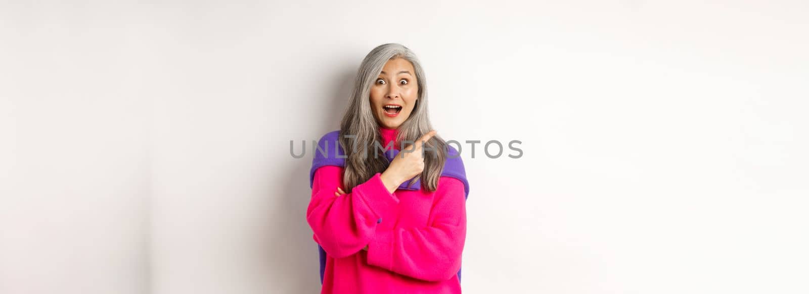 Portrait of impressed asian lady checking out promo offer, pointing at upper left corner and staring at camera amazed, standing in trendy pink sweater over white background by Benzoix