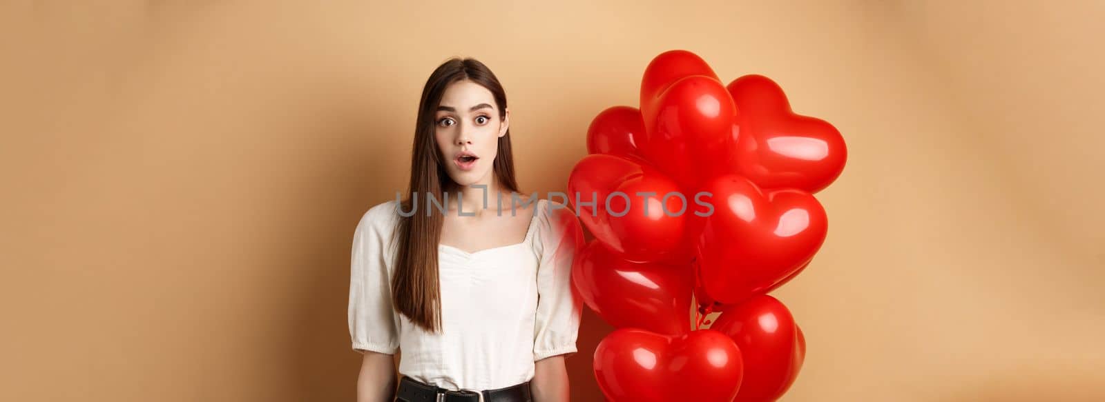 Surprised pretty girl standing near heart balloons and looking with awe at camera. Concept of Valentines day by Benzoix