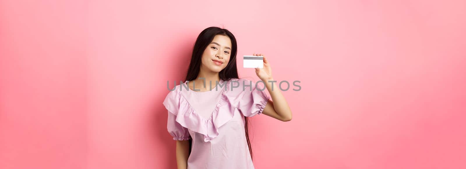 Shopping. Young korean girl in dress showing credit card, smiling satisfied at camera, standing on pink background by Benzoix
