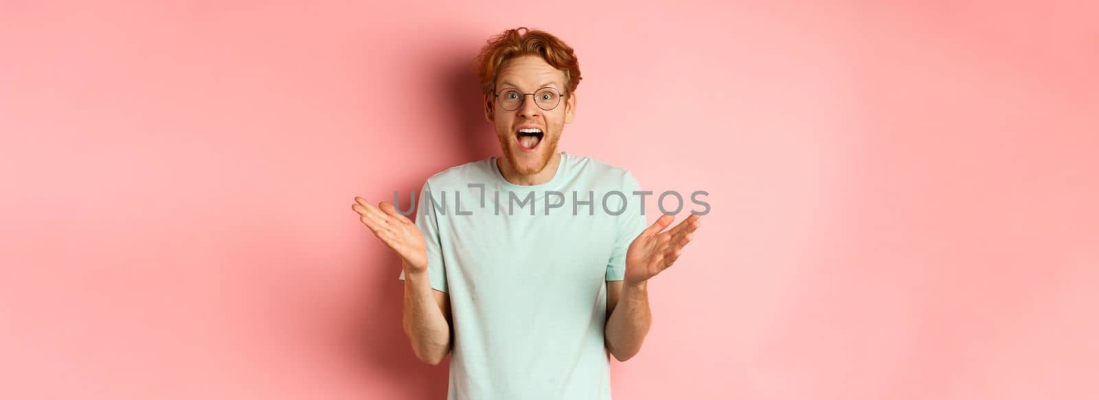 Image of cheerful young bearded man with red hair, wearing glasses, rejoicing of great news, spread hands sideways and congratulating you, standing over pink background.