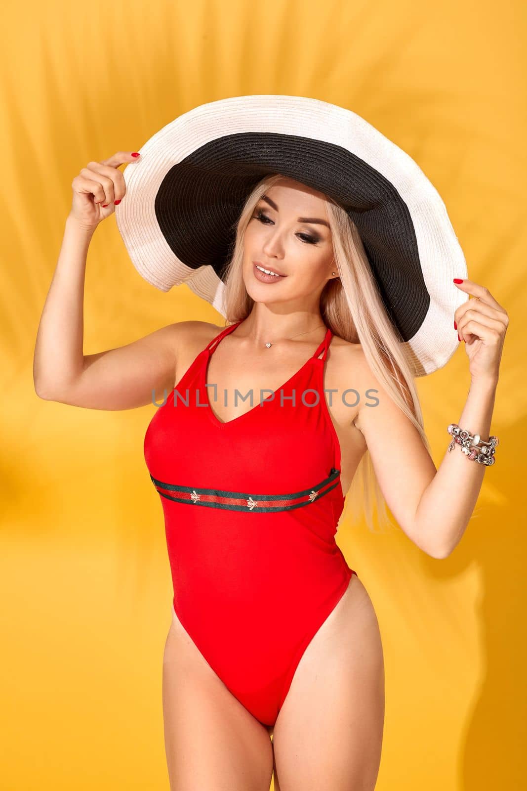 Young sexy blonde woman with long straight hair posing in studio. Perfect gorgeous pale body, red swimsuit or bikini. Big black-and-white hat