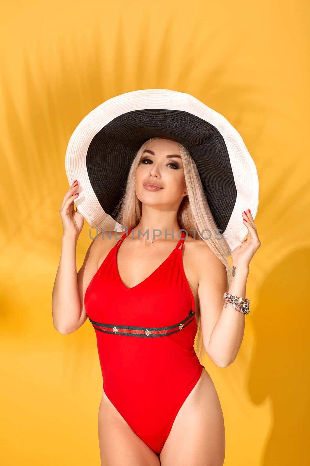 Young sexy blonde woman with long straight hair posing in studio. Perfect gorgeous pale body, red swimsuit or bikini. Big black-and-white hat