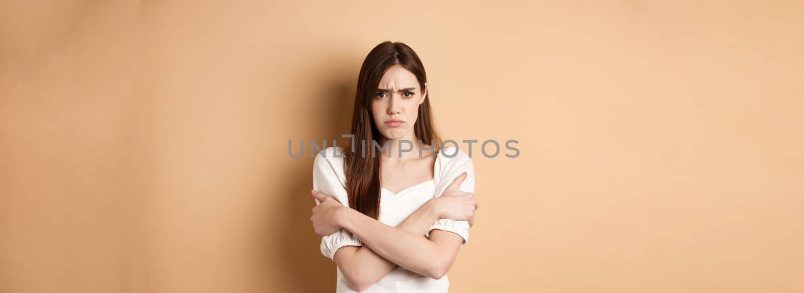 Offended woman hugging herself and frowning upset, feeling timid and defensive, standing on beige background.