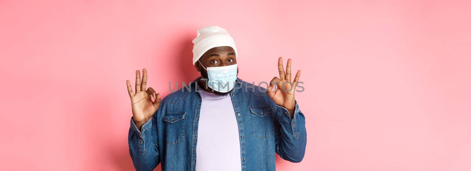 Coronavirus, lifestyle and social distancing concept. Impressed african-american man in face mask, showing okay sign, guarantee and praise, standing over pink background.