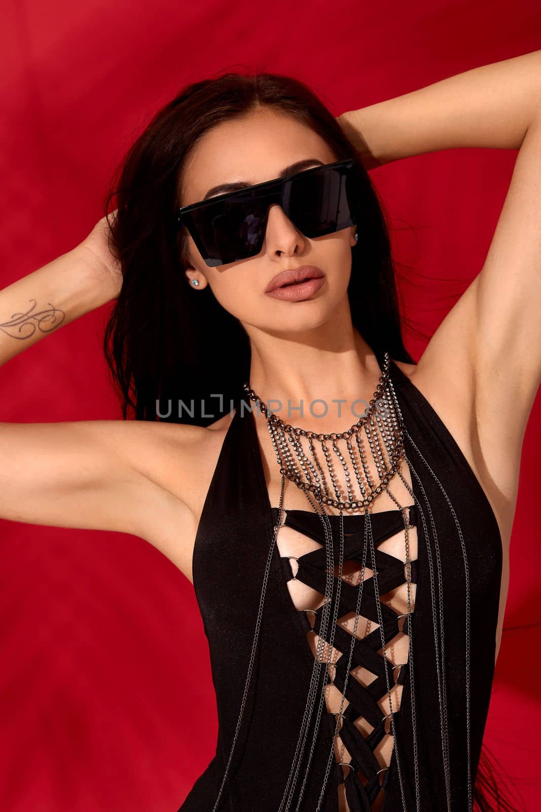 Close up photo of confident attractive young and beautiful woman with long hair in a black sexy swimsuit posing on red background, keeps her arms raised