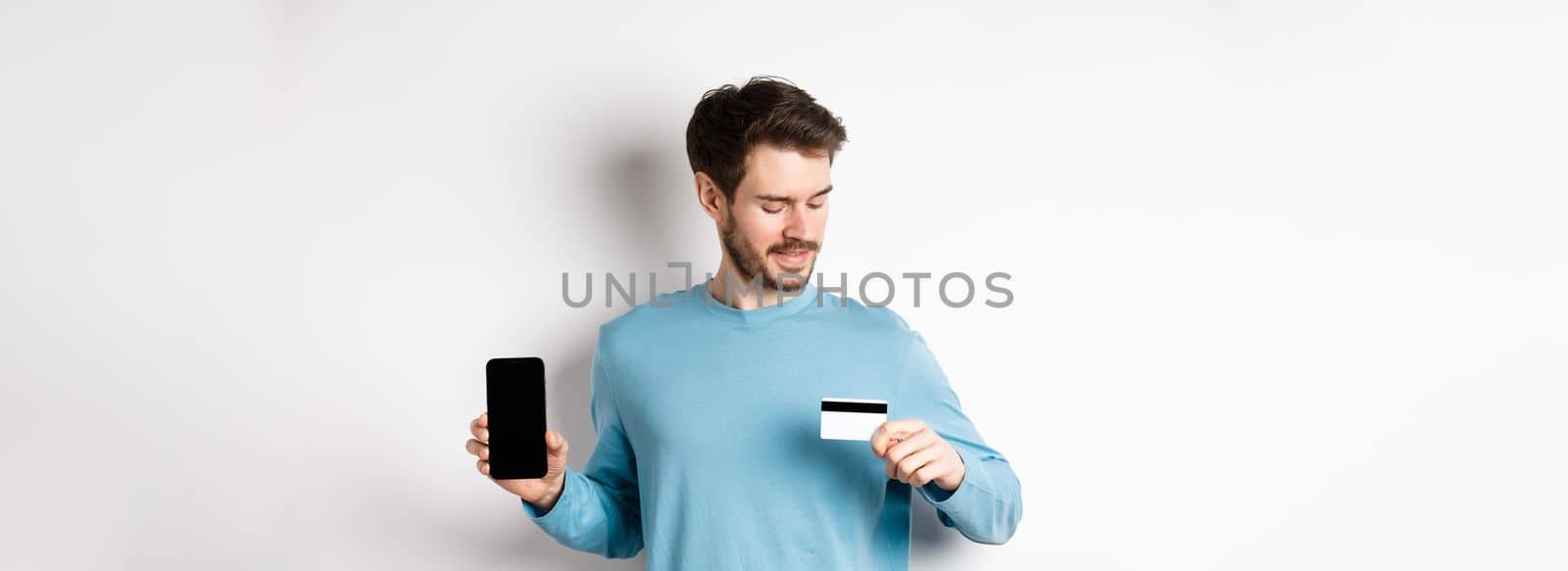 Handsome young man with beard, showing empty smartphone screen and looking at plastic credit card, standing over white background by Benzoix