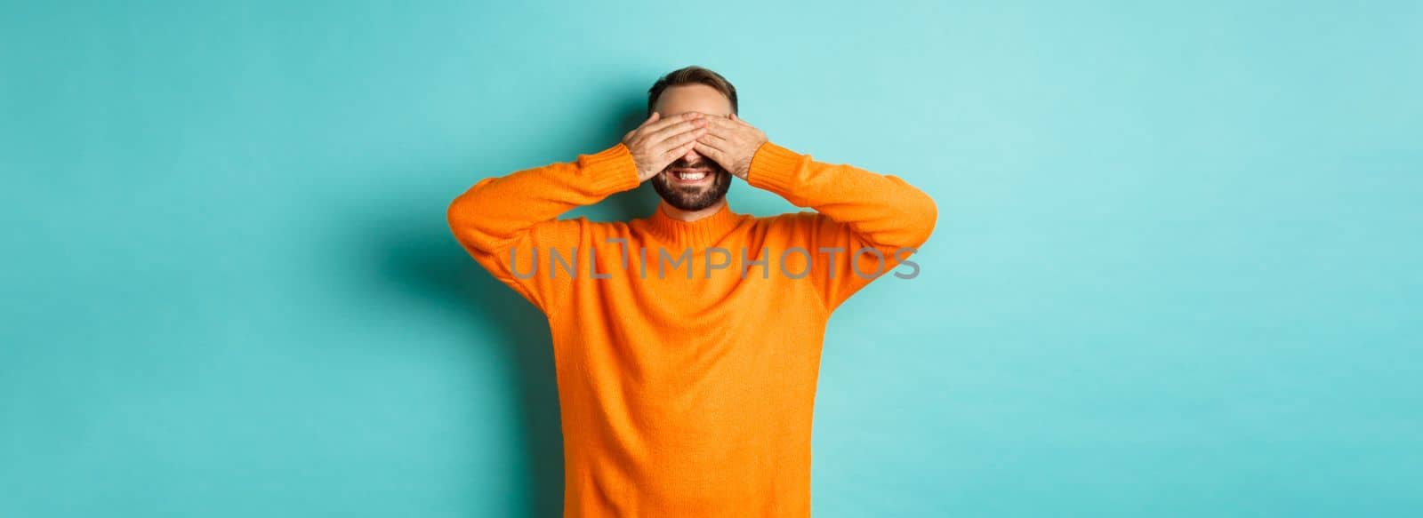 Happy bearded man waiting for surprise, shut ears and expeting gifts, standing over light blue background.