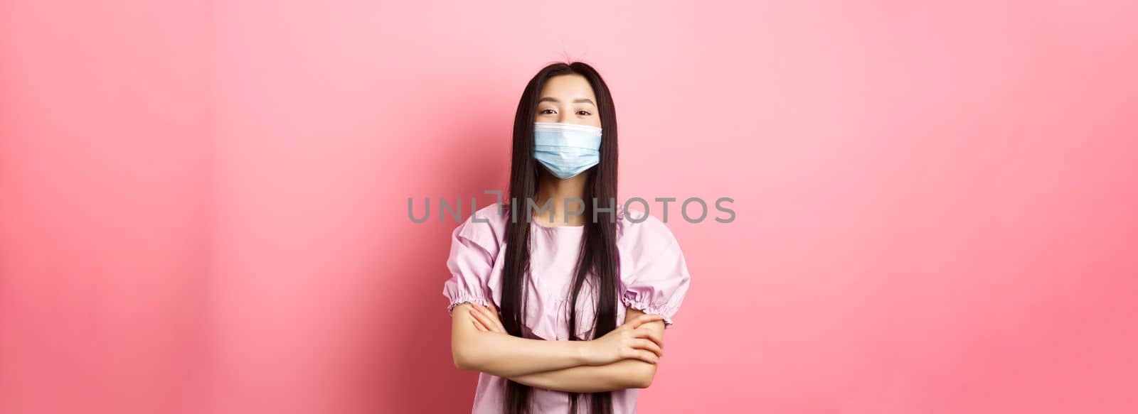 Covid-19, pandemic lifestyle concept. Cute asian woman in medical mask looking confident, cross arms on chest in self-assured pose, standing against pink background.