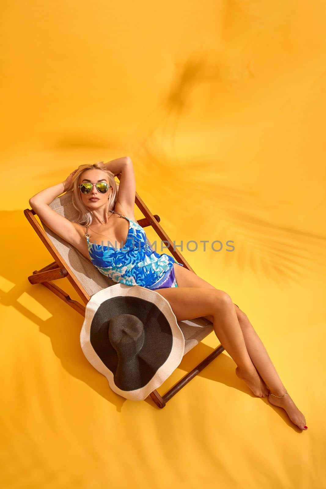 Top view of beautiful young blonde woman in blue swimwear with big sunhat lies on the lounger on yellow background, and taking sunbath