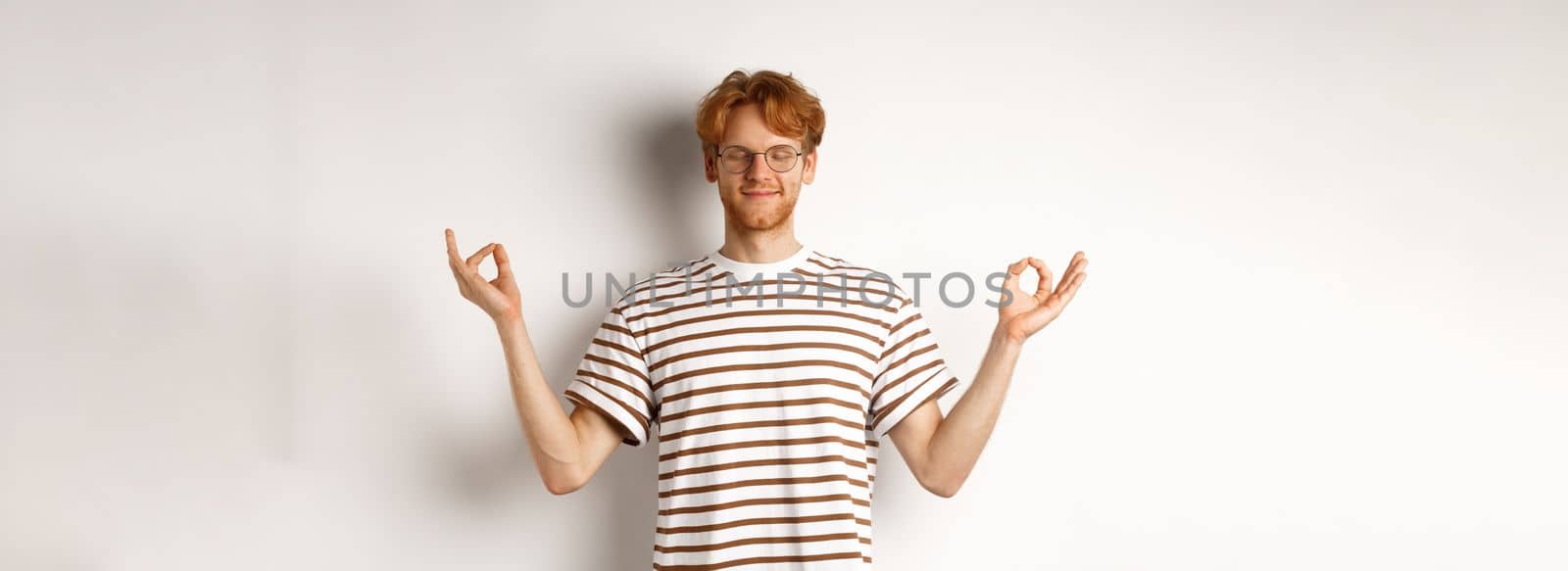 Calm and relaxed young man with red messy hair, spread hands sideways in mudra gesture and smiling, practice yoga or meditating, white background.