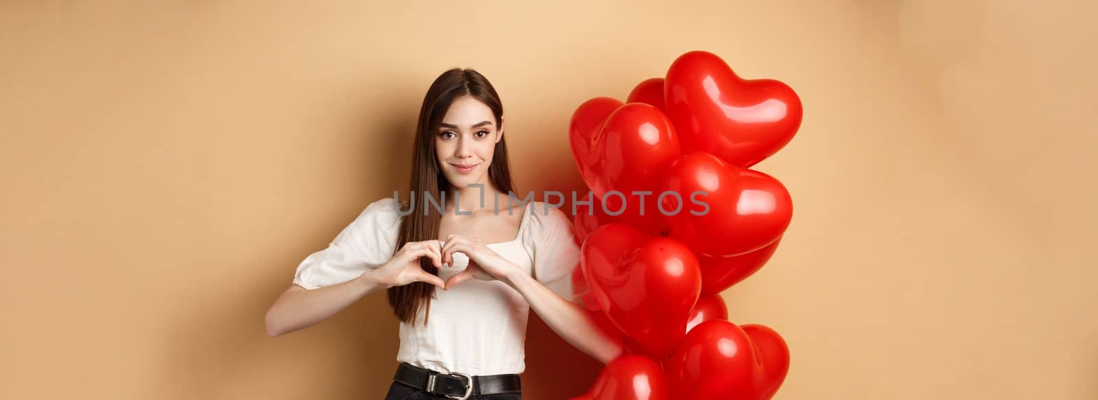 Image of beautiful young woman smiling and showing heart gesture, I love you sign, standing near romantic balloons on Valentines day, beige background by Benzoix