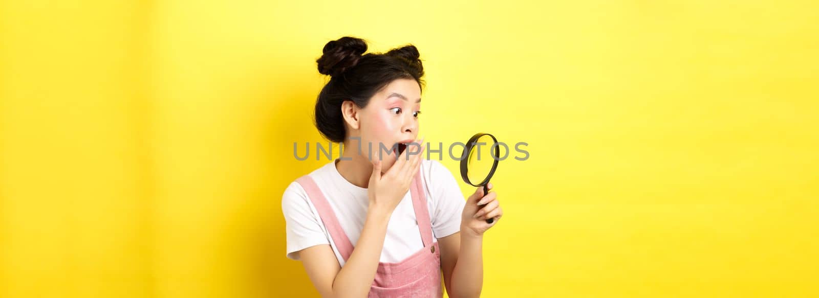 Excited silly asian girl gasping wondered, found something interesting, looking through magnifying glass, standing on yellow background.