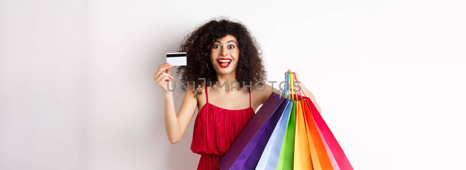 Stylish caucasian woman with curly hair and red dress, showing shopping bags and her plastic credit card, smiling amused, standing over white background by Benzoix