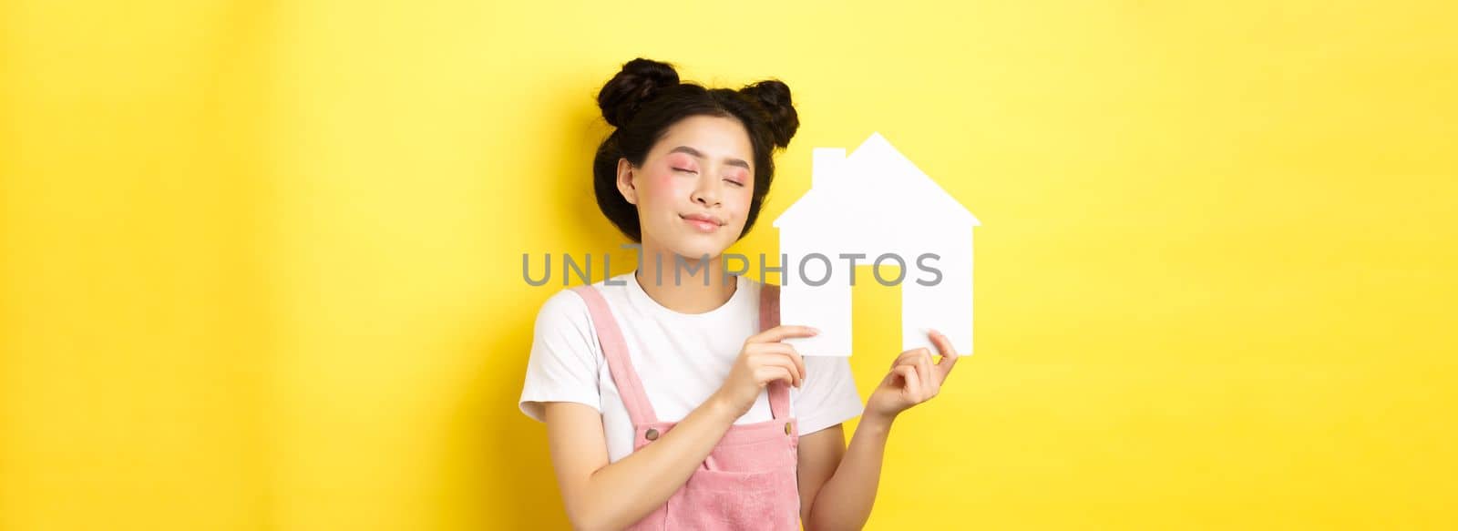 Real estate and family concept. Dreamy smiling asian woman with bright makeup, showing paper house cutout with closed eyes, daydreaming about buying property, yellow background.