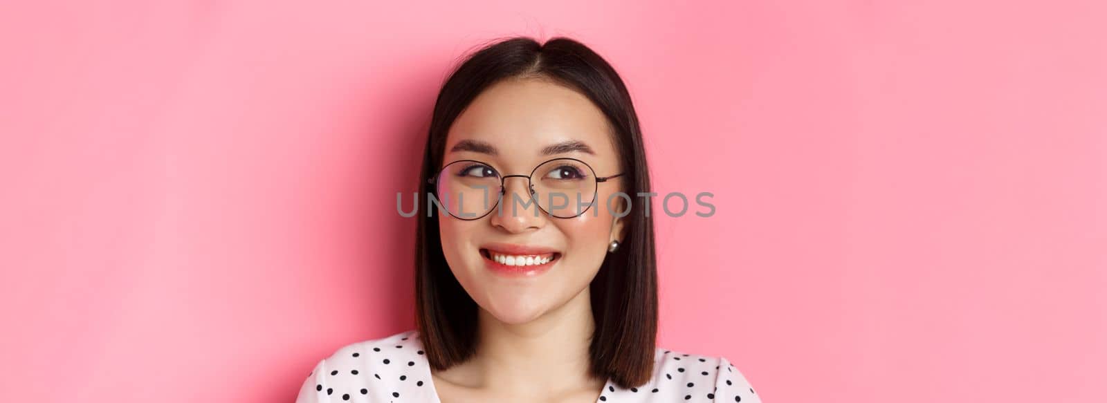 Beauty and lifestyle concept. Close-up of cute asian female model wearing trendy glasses, smiling and looking left at copy space, standing on pink background.