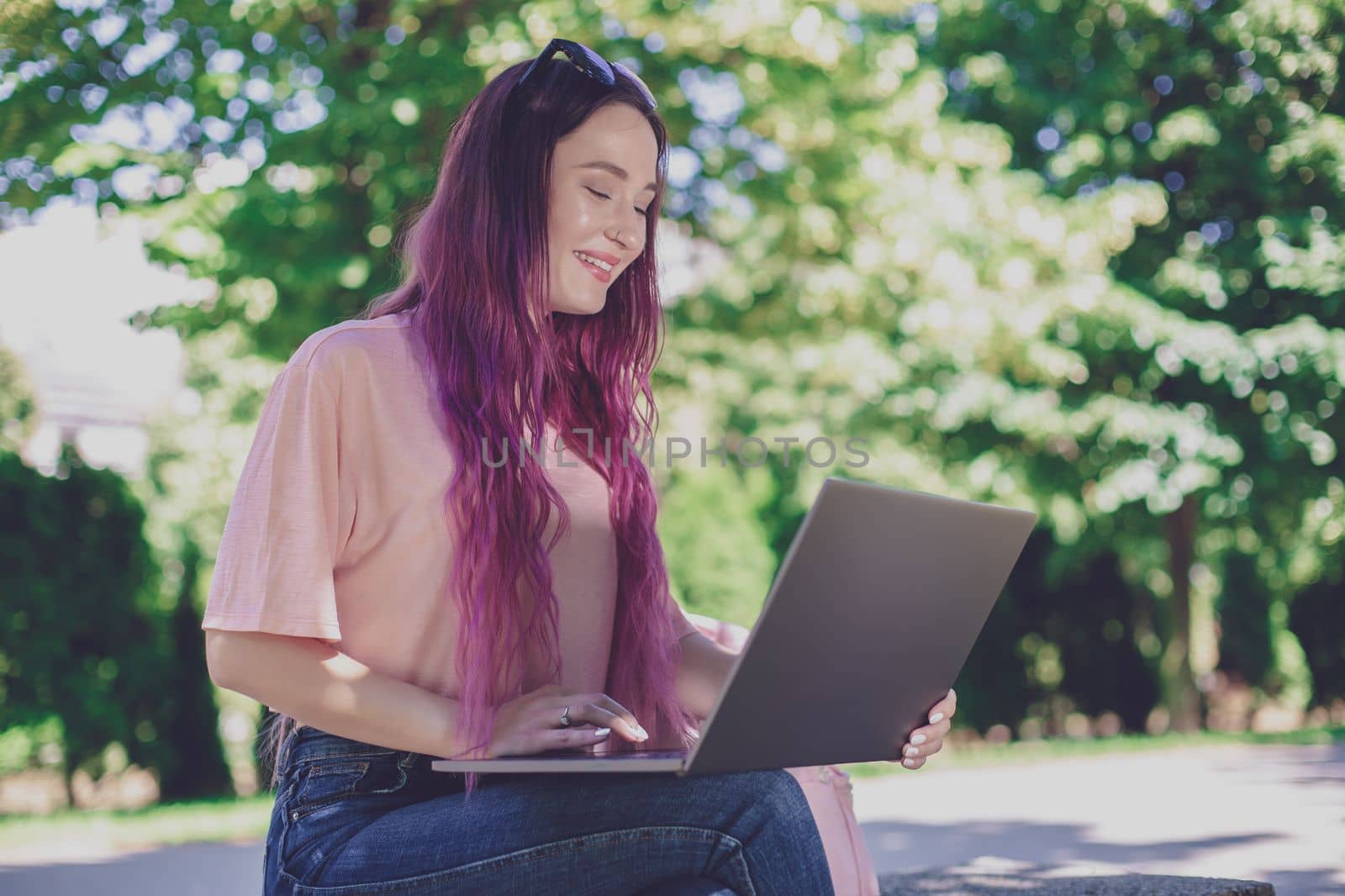 Young girl with pink hair is studying in the spring park, sitting on the wooden bench and browsing on her laptop