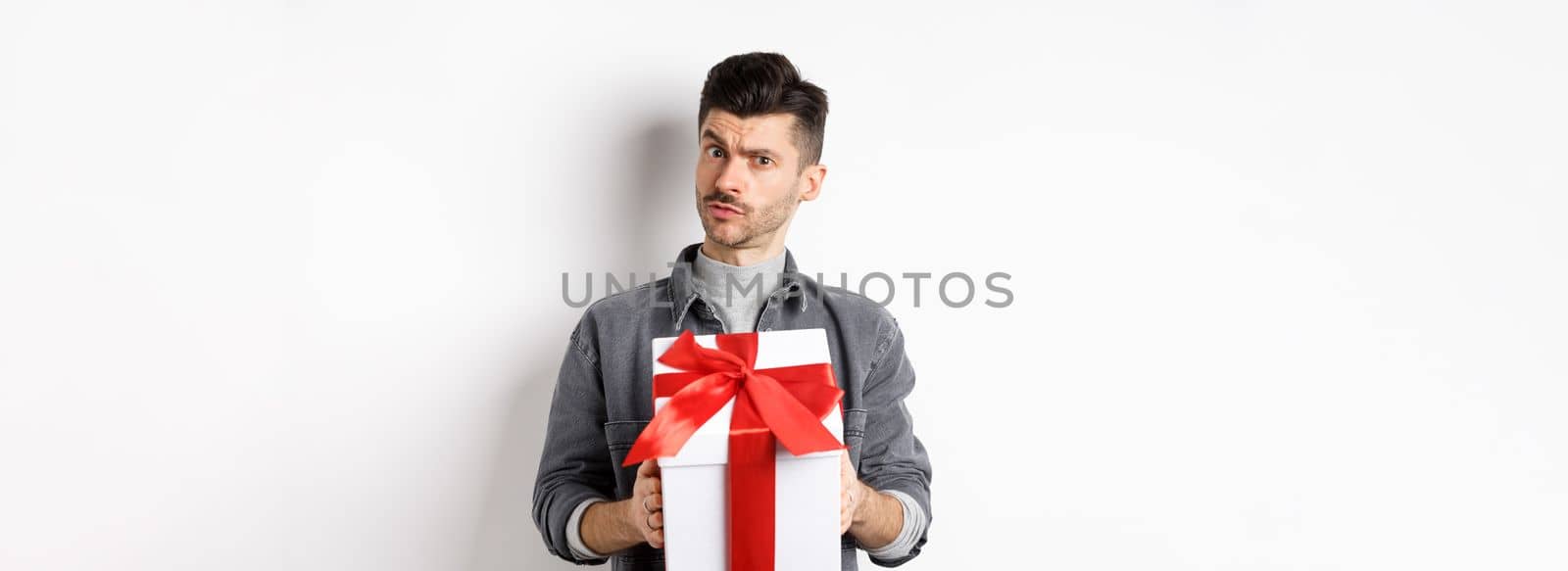 Surprised young man look with disbelief and hold surprise gift, raising eyebrow doubtful, being suspicious, standing on white background. Valentines day concept.