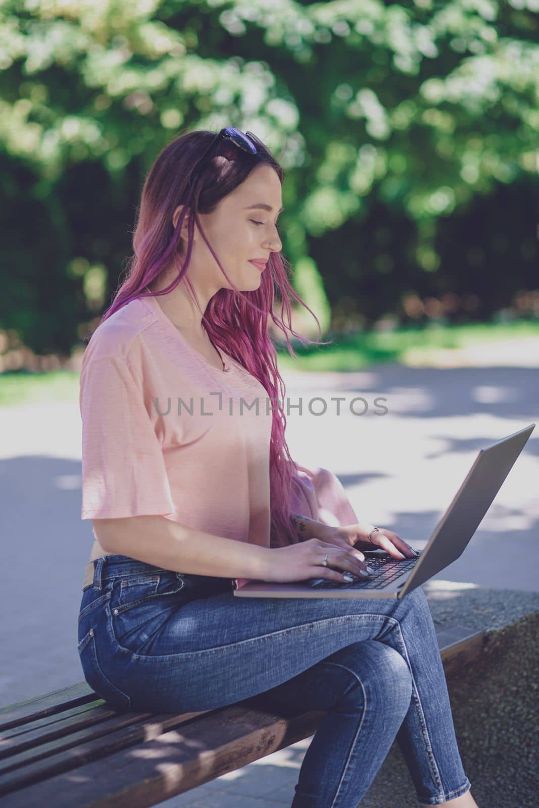 Young girl with pink hair is studying in the spring park, sitting on the wooden bench and browsing on her laptop