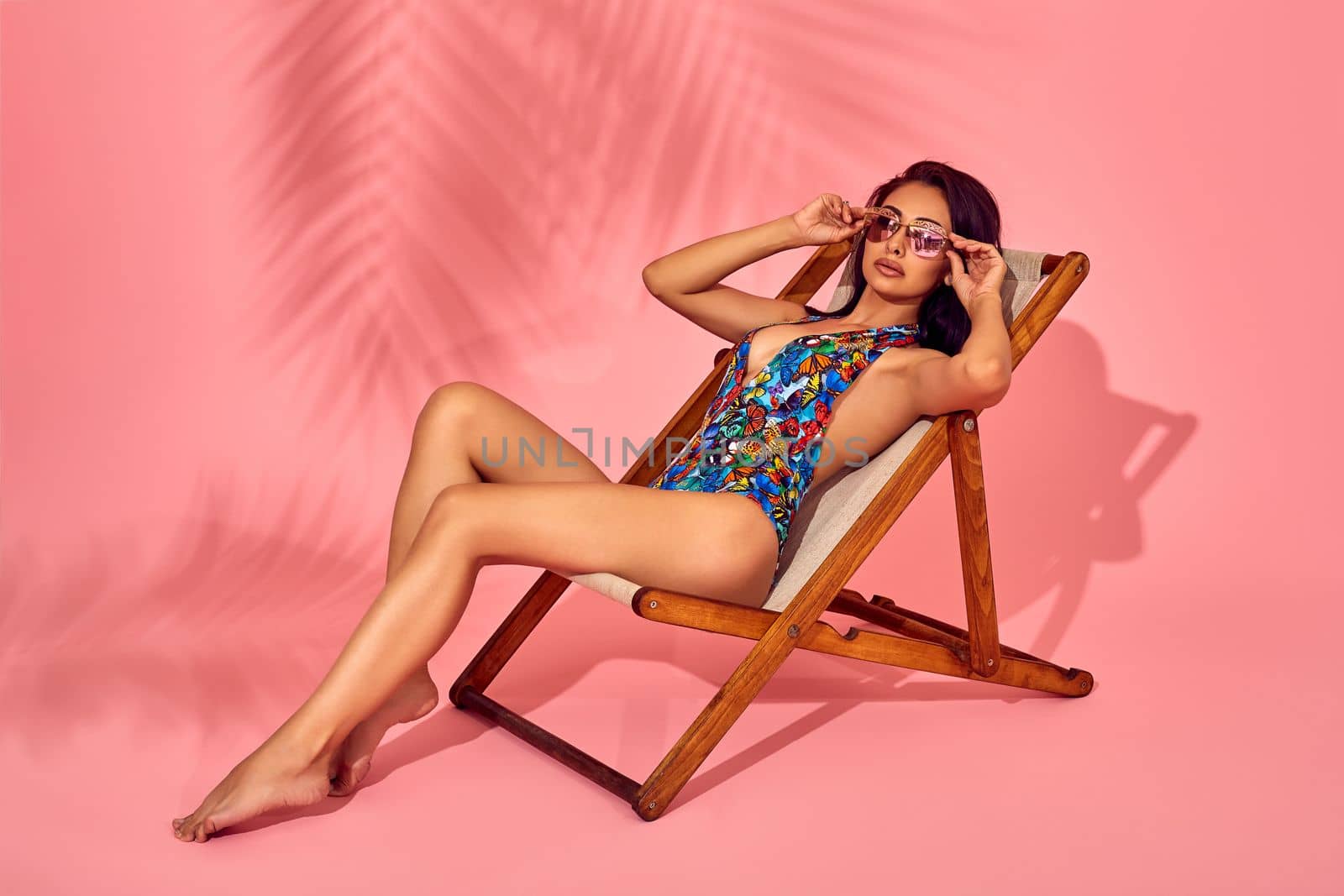 Summer lifestyle fashion portrait of young stunning woman on a lounge chair, pink background, studio shot. Wearing stylish sunglasses, bikini. by nazarovsergey