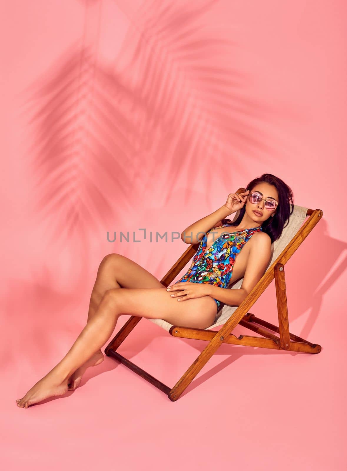 Summer lifestyle fashion portrait of young stunning woman on a lounge chair, pink background, studio shot. Wearing stylish sunglasses, bikini. by nazarovsergey