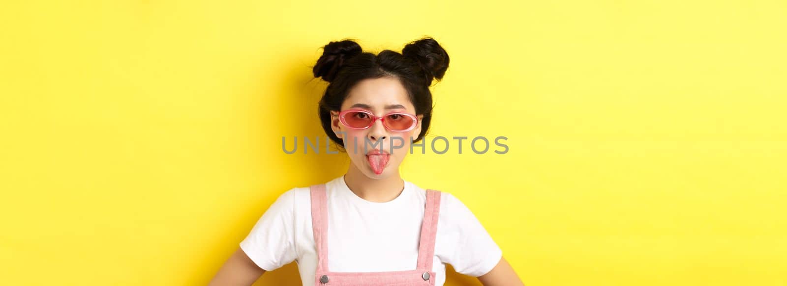 Summer and fashion concept. Silly asian teen girl showing tongue, wearing sunglasses, standing on yellow background by Benzoix