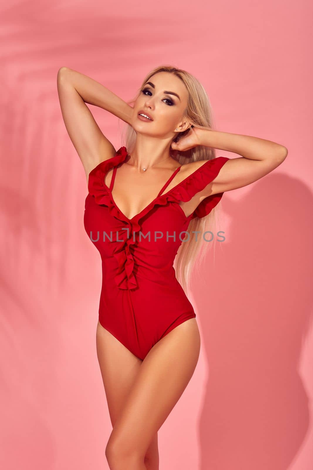 Stunning blonde female model with amazing body standing in an elegant red swimsuit on pink background with palm shadow. Vacation and recreation concept