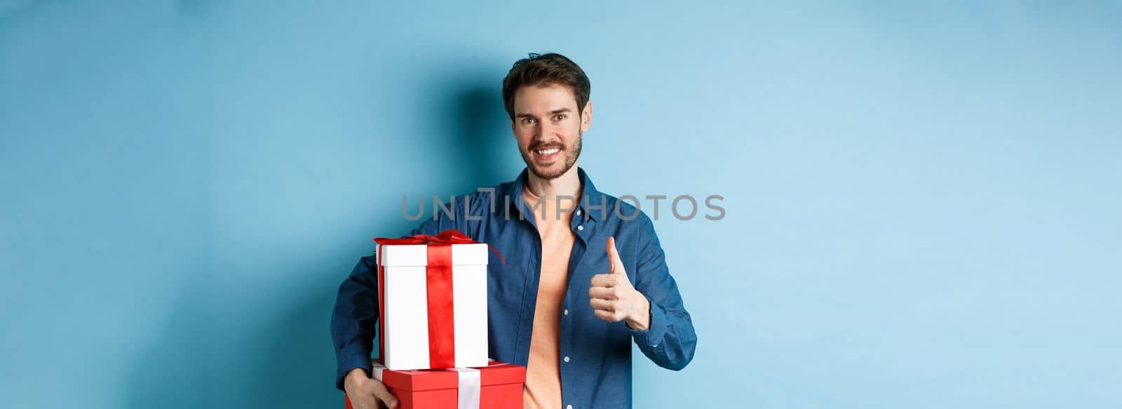 Smiling man holding romantic gifts and showing thumbs-up, celebrating Valentines day, buying presents for lover, standing over blue background.