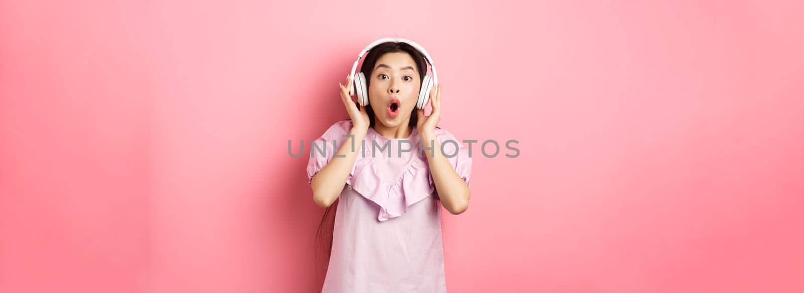 Excited asian girl hear awesome song, listen music in headphones and look amazed, standing on pink background by Benzoix