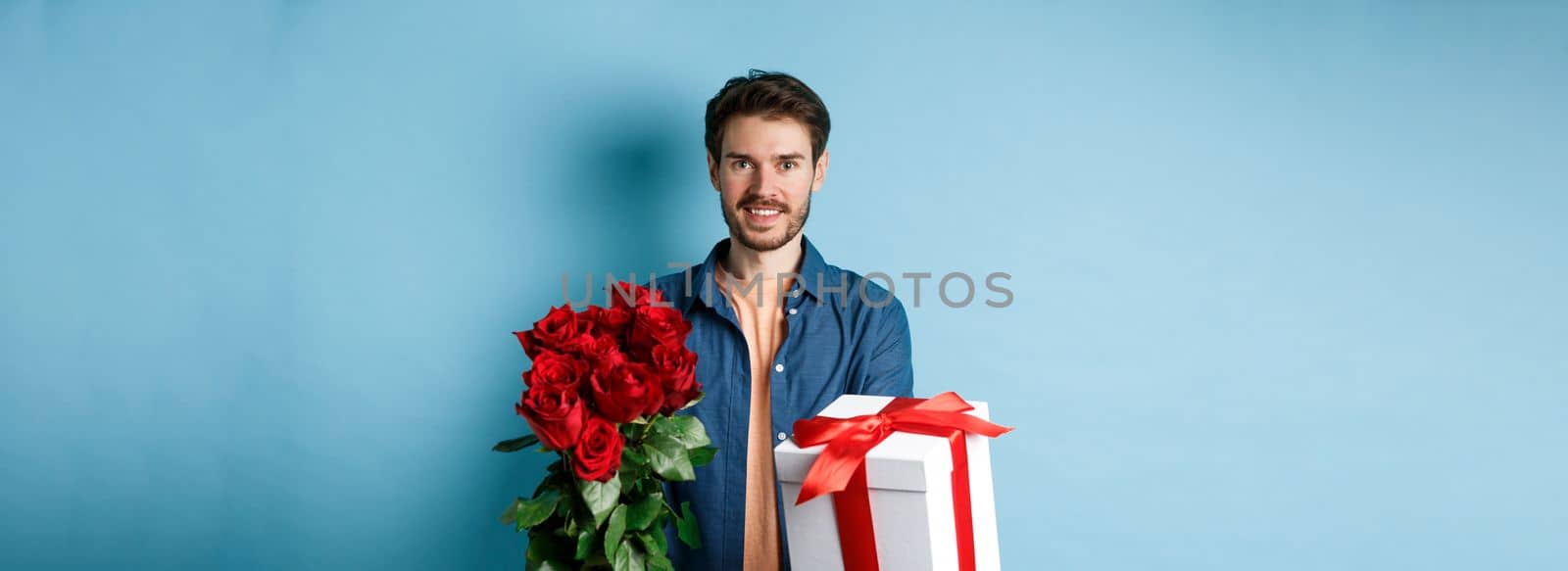 Love and Valentines day concept. Charming young man giving gift and bouquet of roses to girlfriend, standing over blue background.