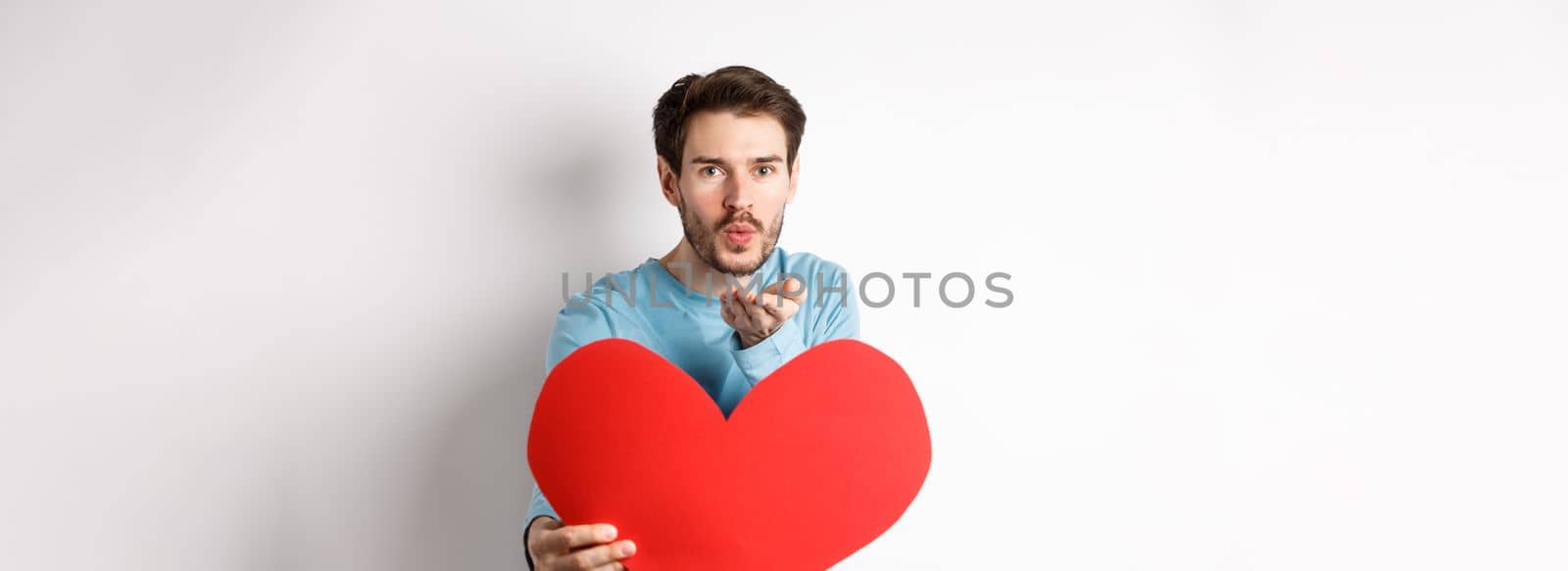Romantic boyfriend sending air kiss at camera and holding big red Valentines day heart, standing over white background.