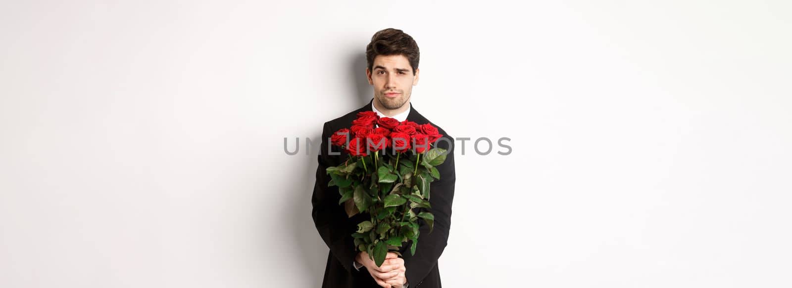 Image of handsome boyfriend in black suit, holding bouquet of red roses and smiling, being on a date, standing over white background.