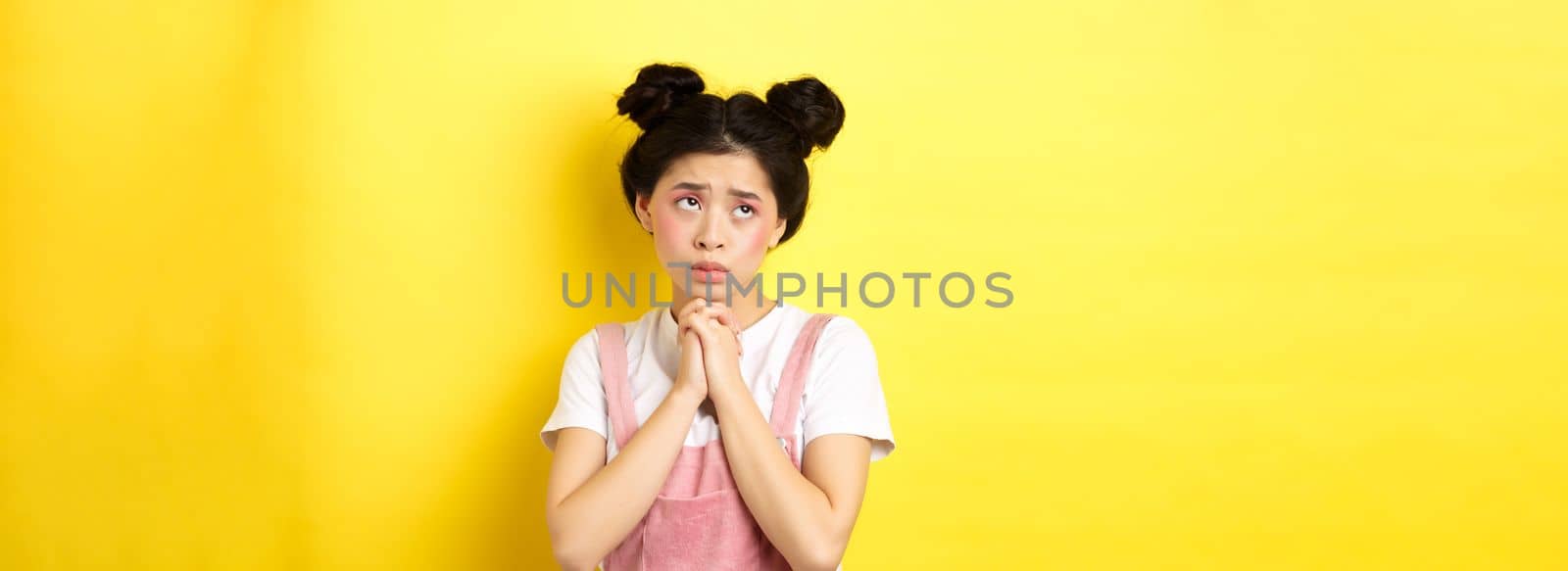 Sas asian girl holding hands in pray, pleading god, looking up and begging, standing on yellow background.