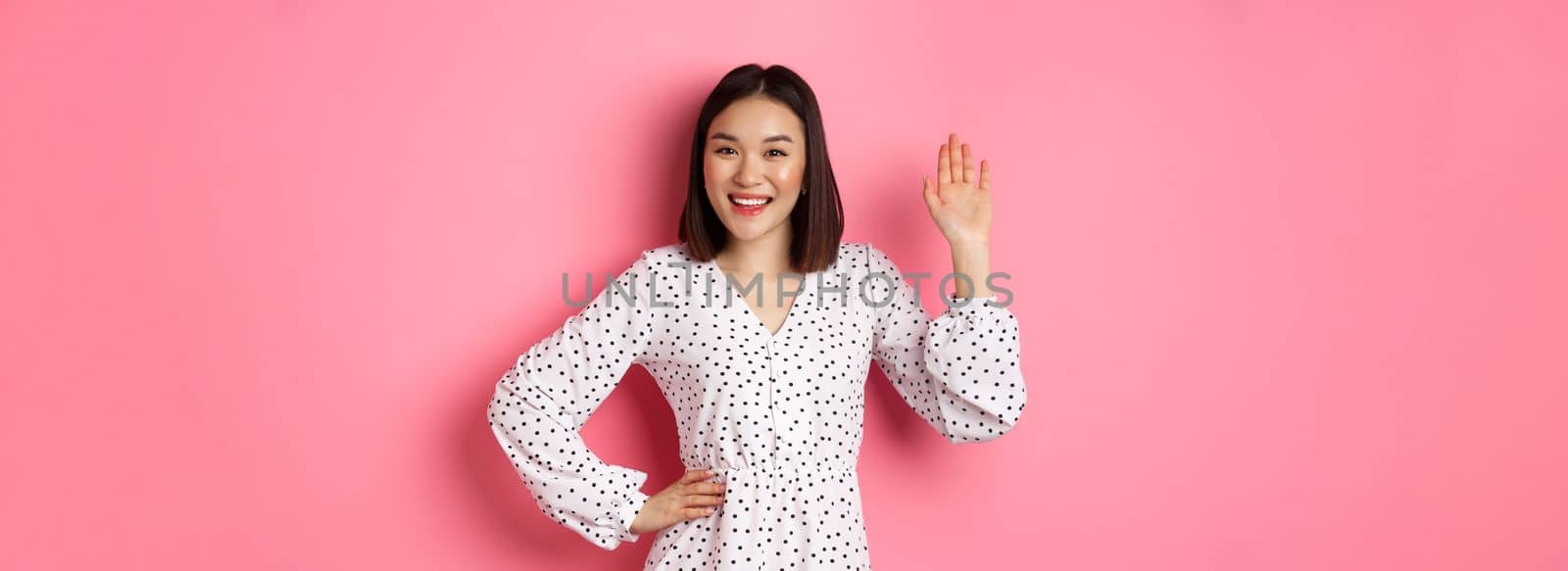 Beautiful asian woman in dress saying hello, waving hand to greet and say hi, smiling friendly at camera, standing over pink background by Benzoix