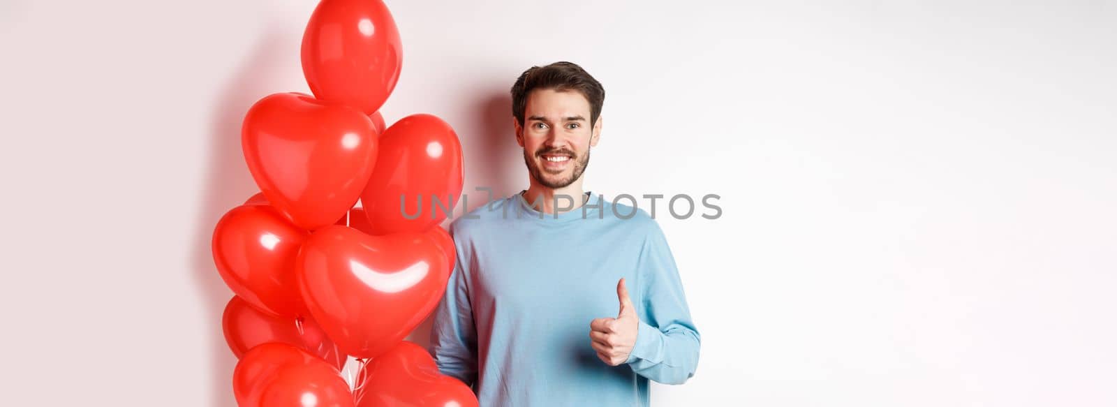 Valentines day and lovers concept. Happy young man showing thumbs up as standing with red hearts balloon, bring romantic gift on date, standing over white background by Benzoix