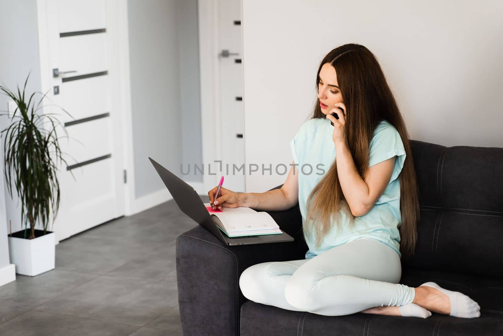 Busy girl with laptop working online and talking with colleagues by phone at home. Young business woman is sitting on sofa and thinking about creative idea of new project and noticing best ideas