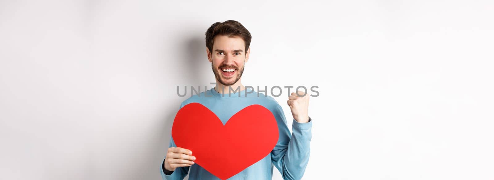 Cheerful man determined to find love on Valentines day, making fist pump success gesture and holding big red romantic heart, standing over white background by Benzoix