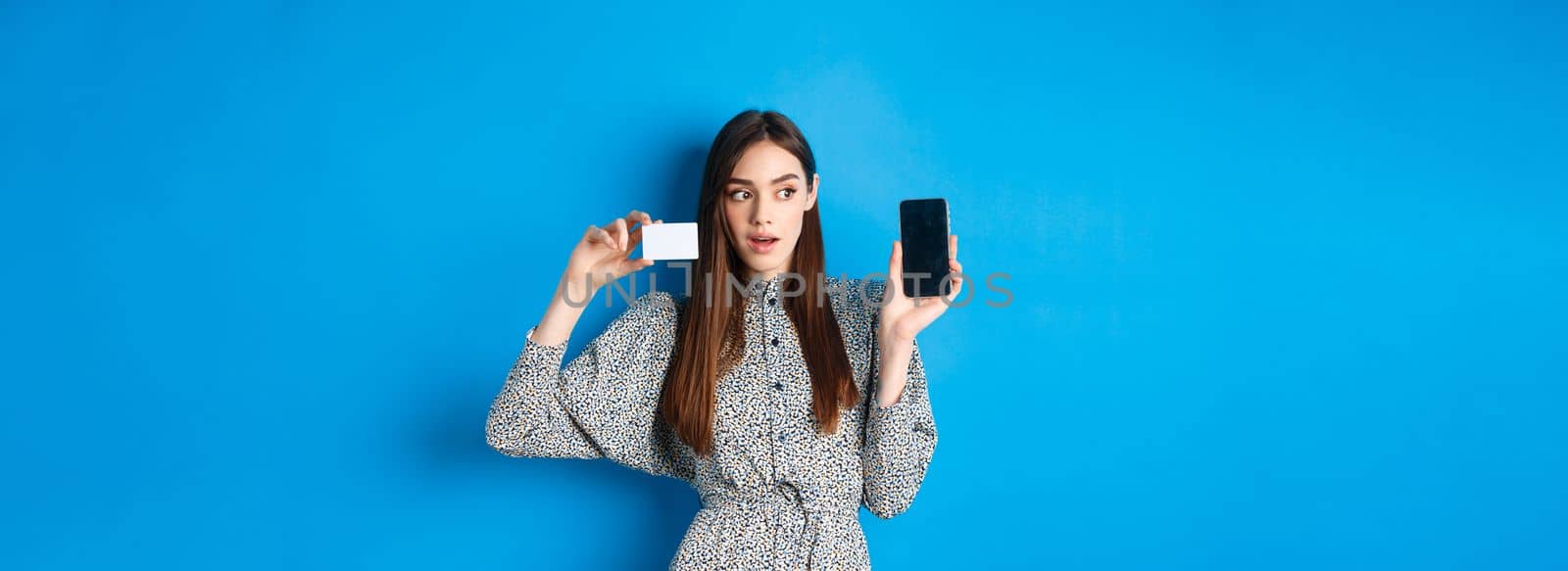 Online shopping. Pensive lady in dress showing empty phone screen and plastic credit card, order in internet, blue background.