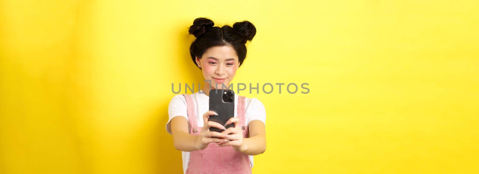 Stylish asian girl taking photo on smartphone, making video with cellphone and smiling, standing on yellow background.