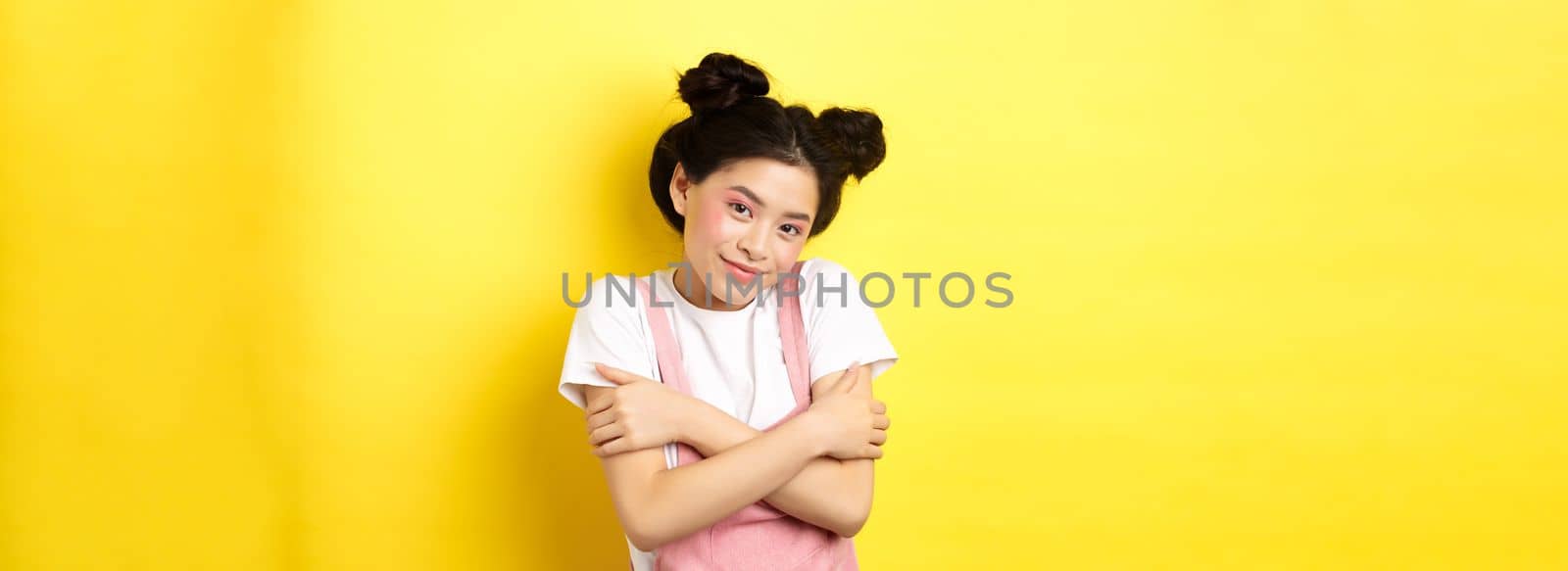 Summer lifestyle concept. Cute asian beauty girl with makeup, hugging herself and smiling romantic, standing tender on yellow background.