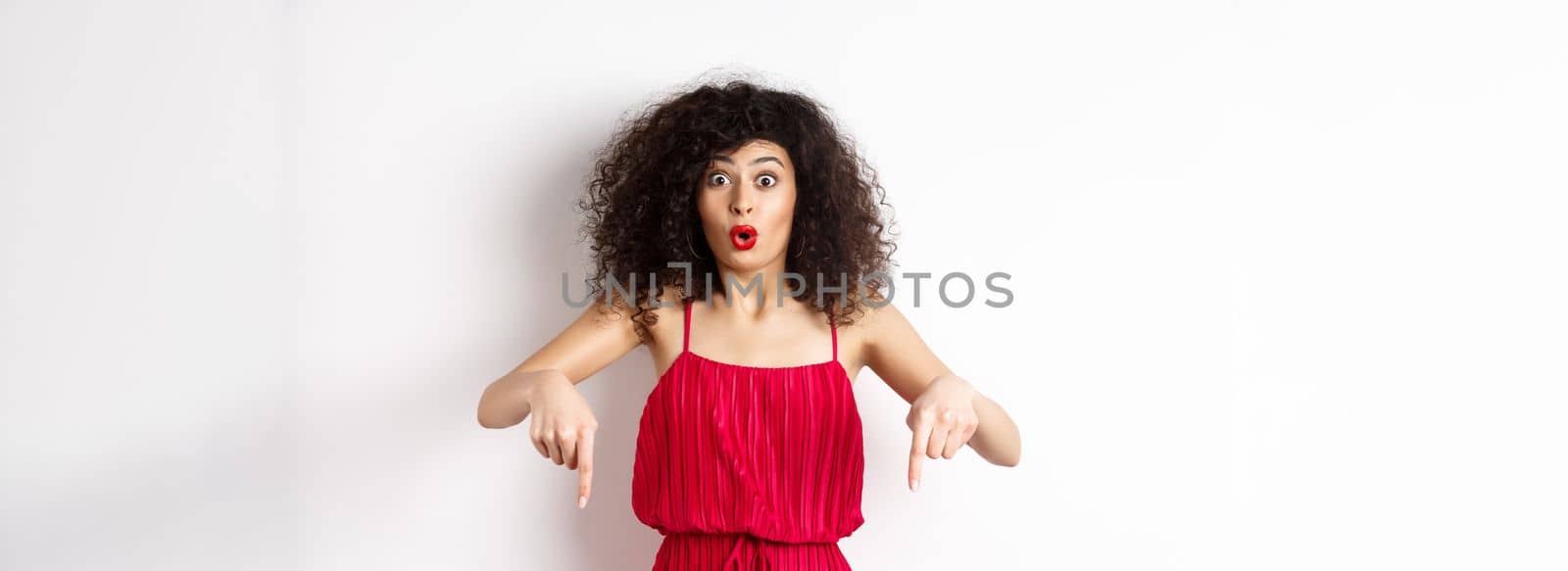 Impressed woman in red dress say wow, showing promo deal, pointing fingers down with amazed face, standing over white background.