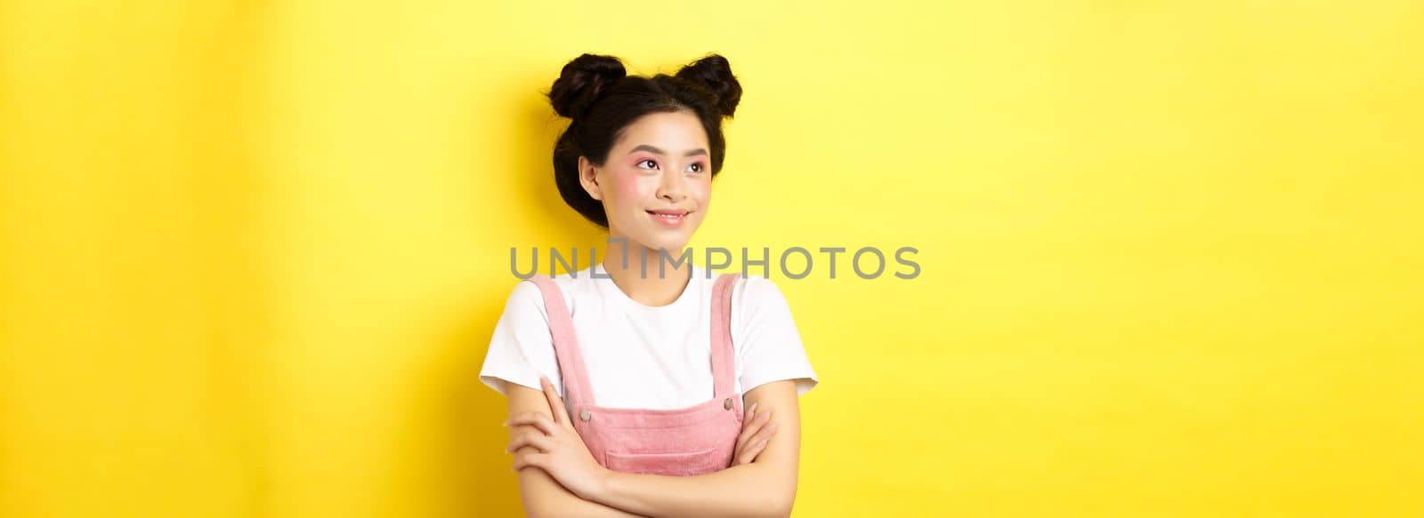 Smiling beautiful asian girl with glamour makeup, looking aside at logo with happy face, standing in summer clothes on yellow background.