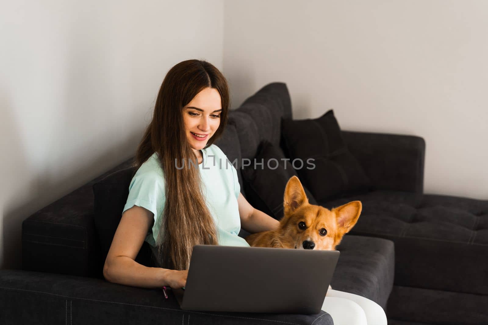 Attractive girl hug Corgi dog and have a rest at home. Girl and her domestic pet sit on the sofa and watching film on laptop together. Lifestyle with Welsh Corgi Pembroke. by Rabizo