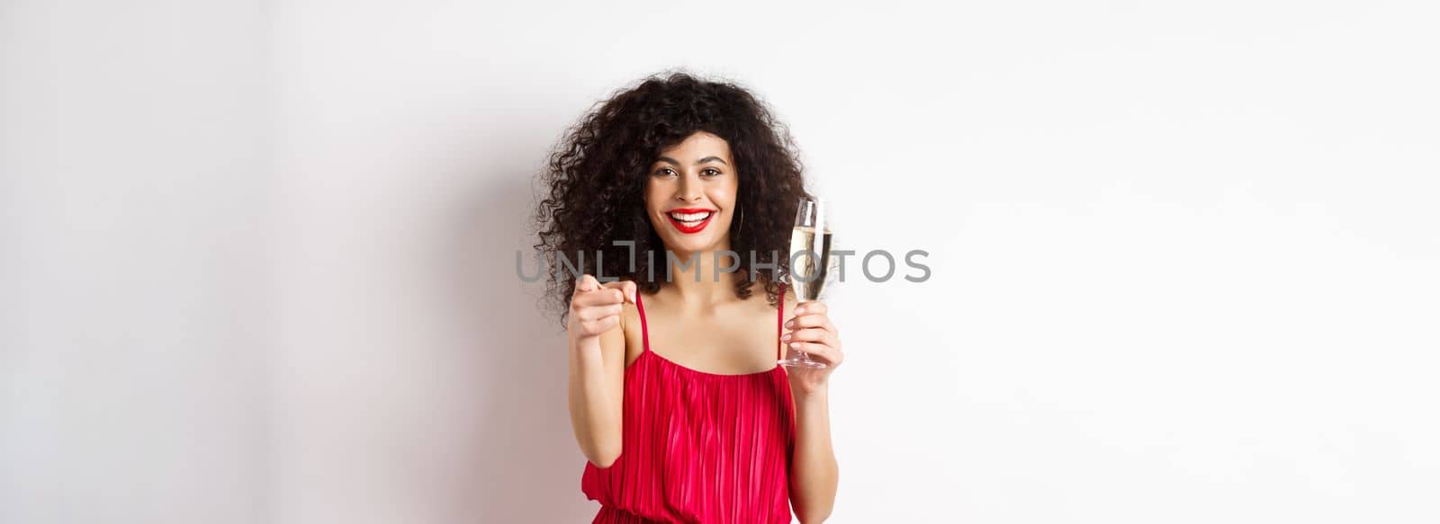 Beautiful curly woman in red dress, partying or having romantic date, holding glass of champagne and pointing at you, inviting person, standing on white background.