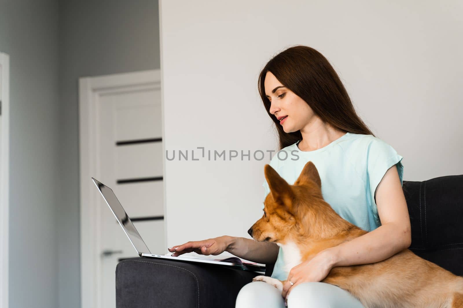 Attractive girl hug Corgi dog and have a rest at home. Girl and her domestic pet sit on the sofa and watching film on laptop together. Lifestyle with Welsh Corgi Pembroke